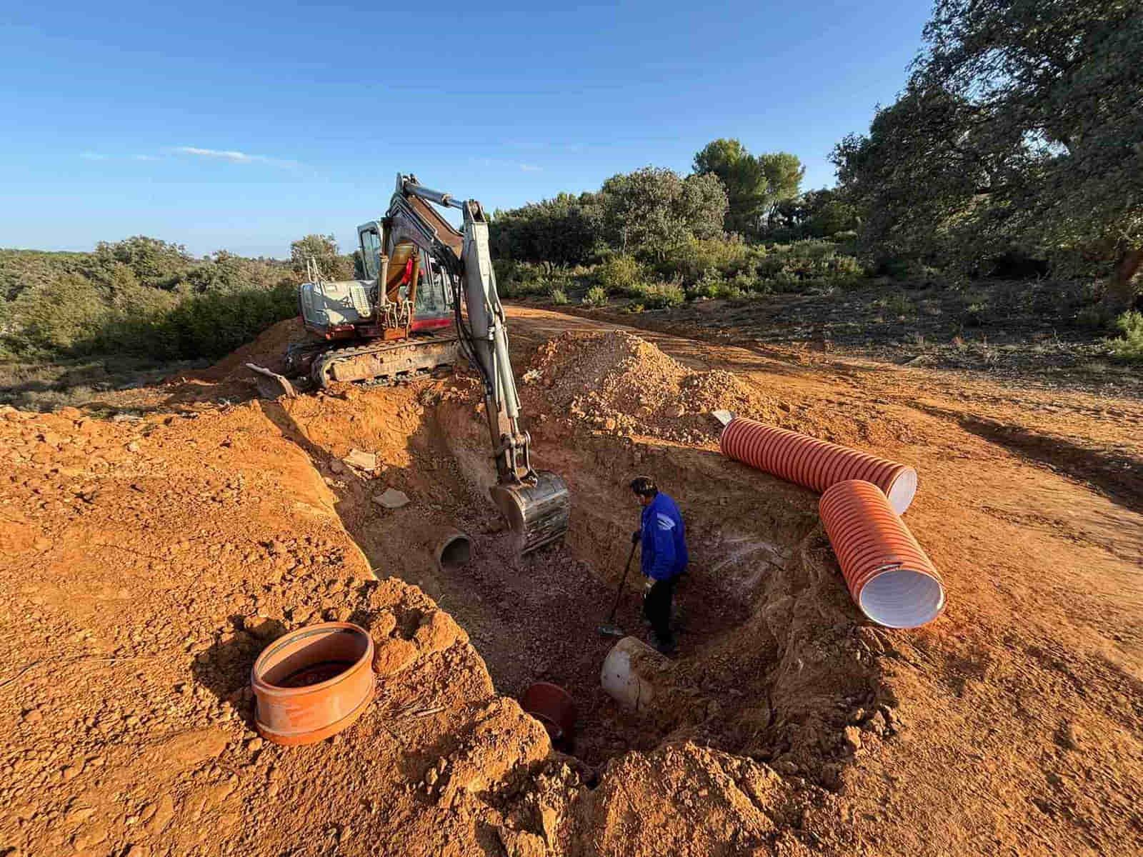 Excavadora cavando zanja para tuberías en un camino de tierra; trabajador con camisa azul cerca de la zanja; tubos naranjas.