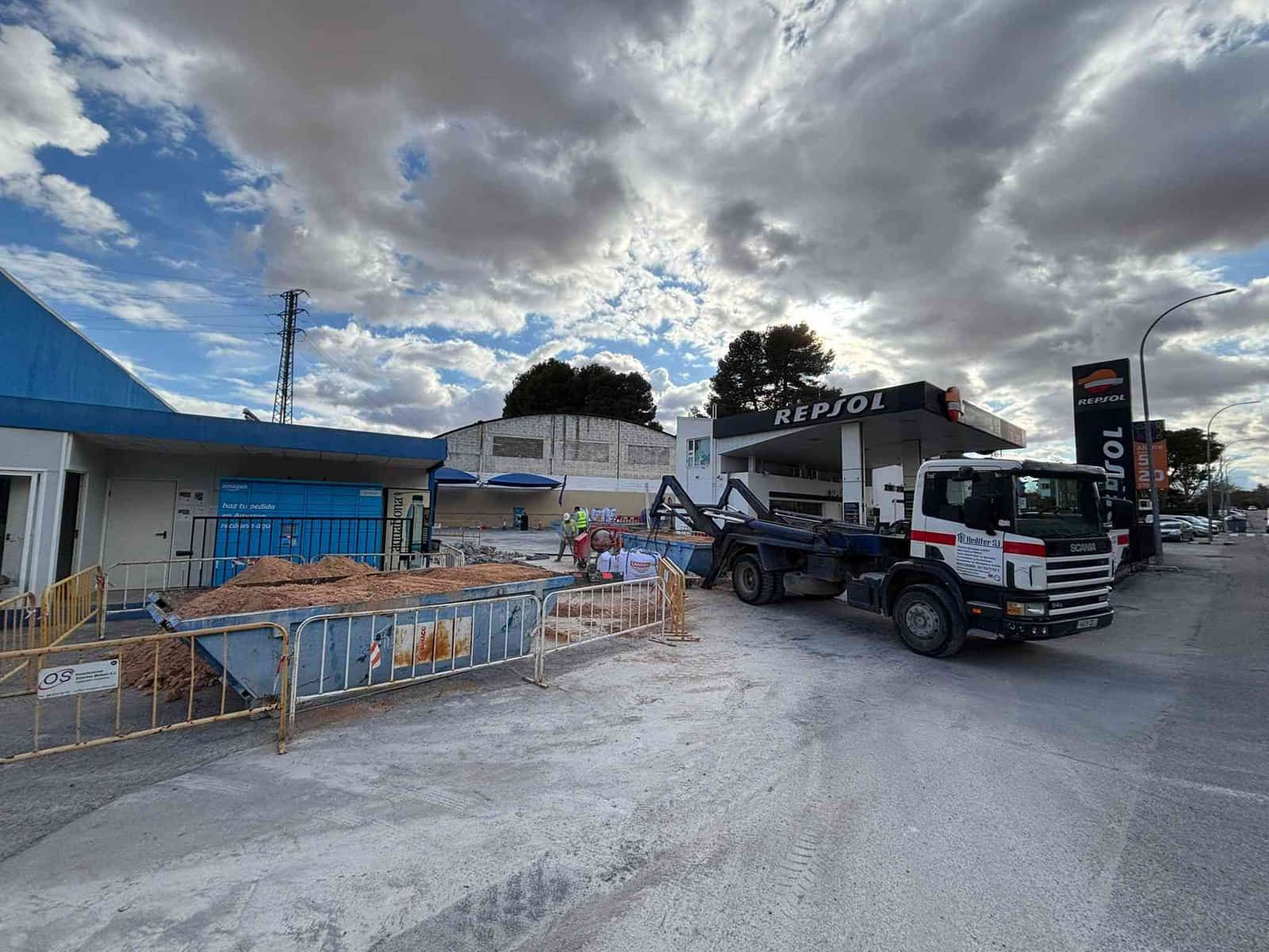 Obras en gasolinera Repsol: obreros, camión y excavadora visibles bajo un cielo nublado.
