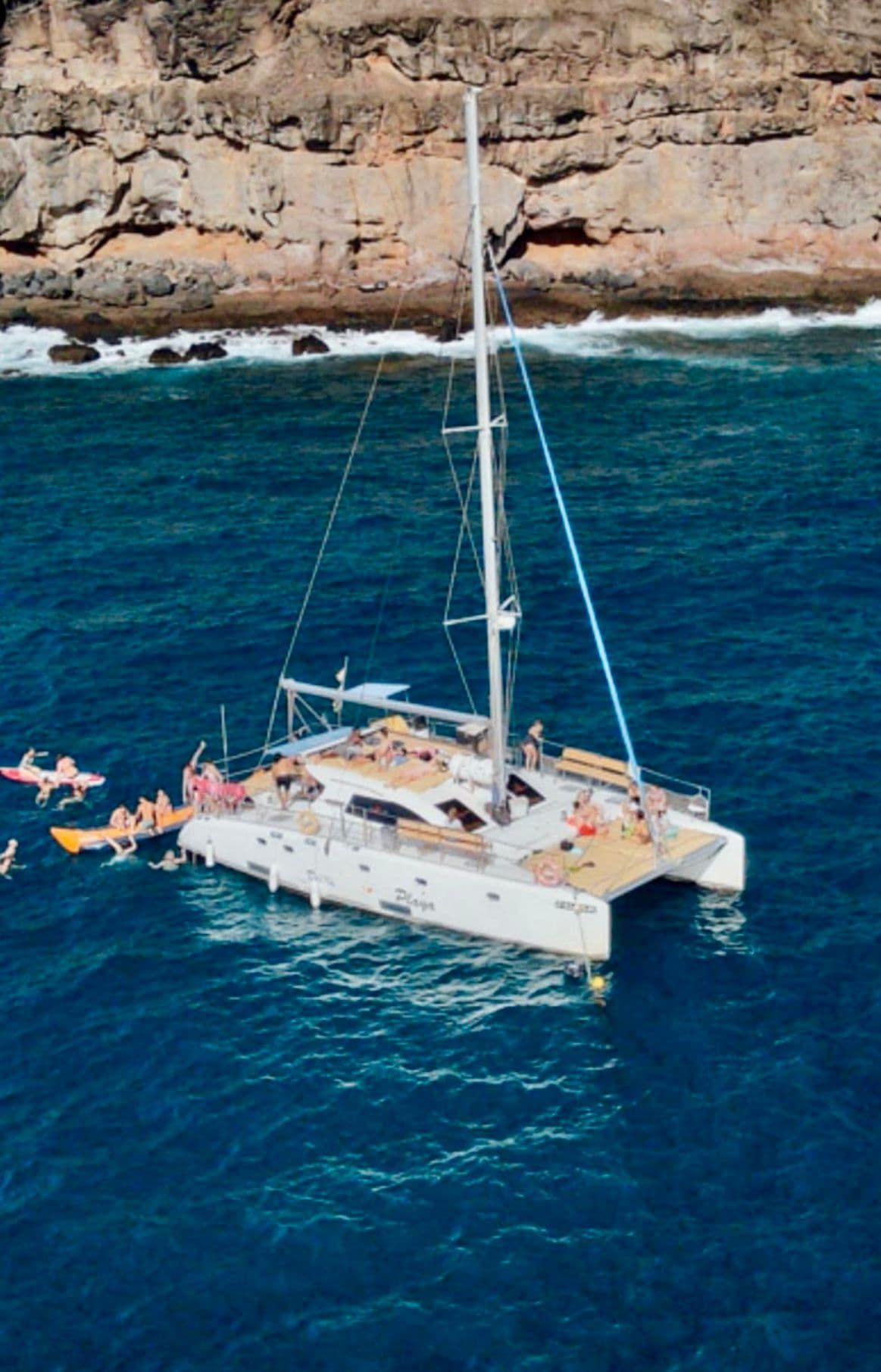 Catamarán navegando en aguas azules cerca de un acantilado rocoso, con gente nadando y relajándose a bordo.