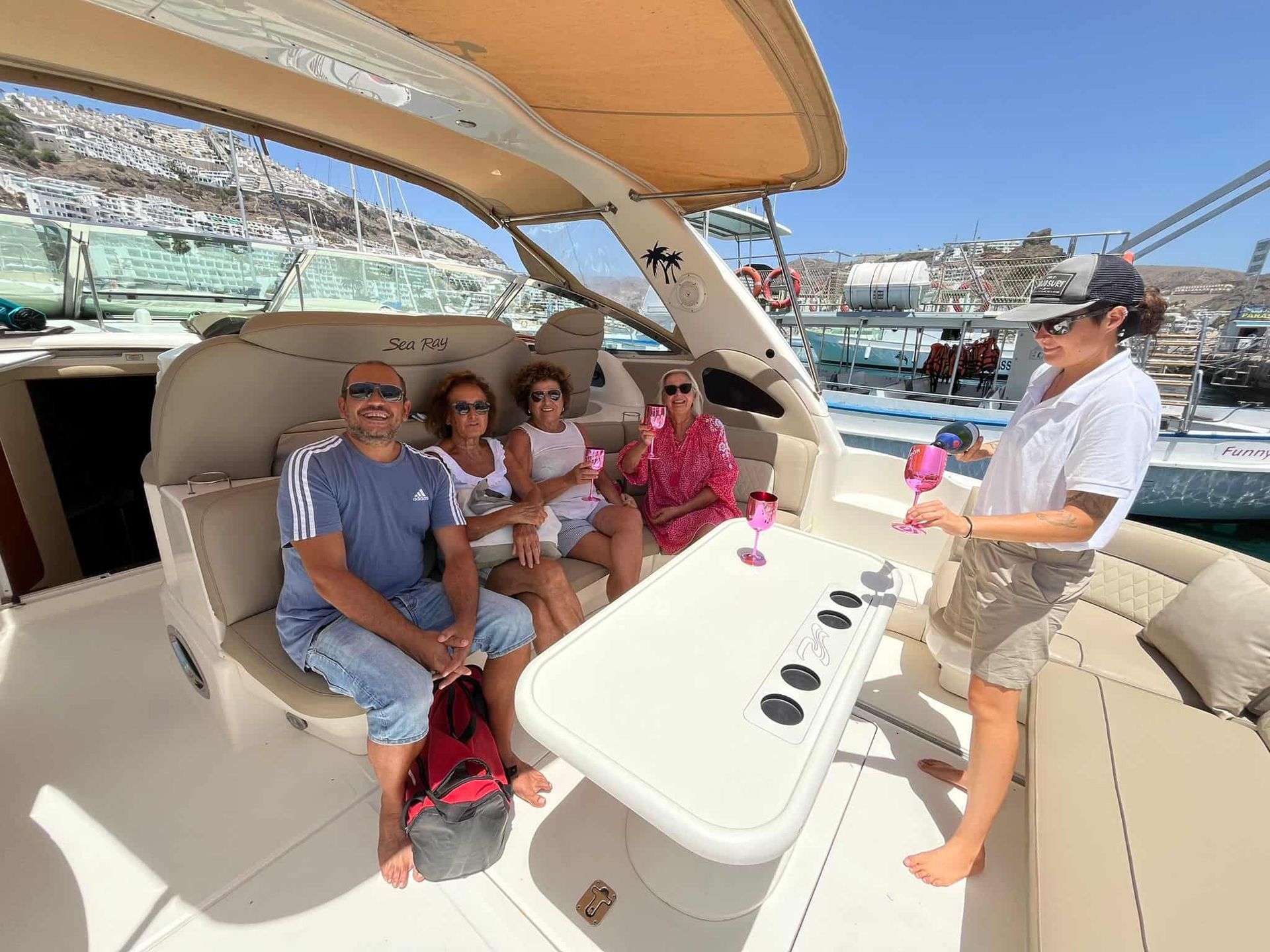 Cuatro personas en un barco con bebidas. Una persona sirve una bebida. Día soleado, vista del puerto.