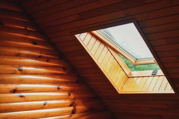 In einem rustikalen Zimmer befindet sich ein Oberlichtfenster in einer holzgetäfelten, schrägen Decke neben einer geschwungenen Blockwand.