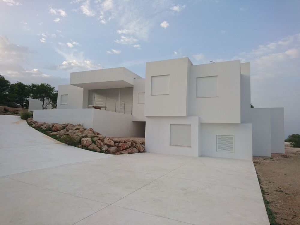 Edificio blanco moderno con formas geométricas apiladas sobre un camino de hormigón, contra un cielo azul claro.
