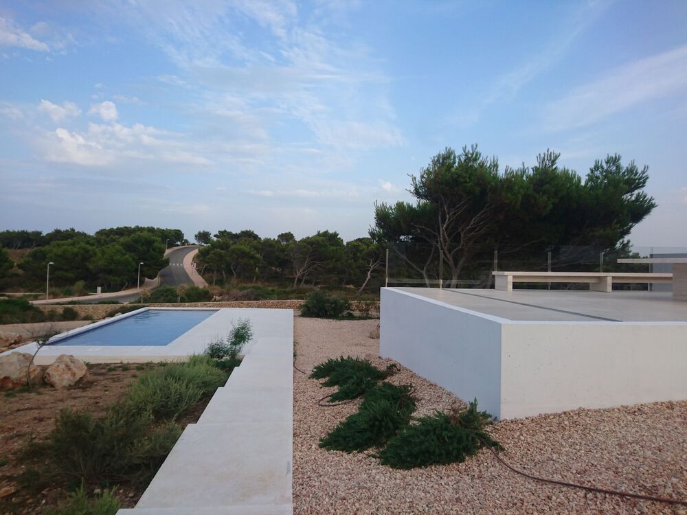 Piscina y moderno edificio blanco en un entorno natural, con vistas a una carretera y árboles bajo un cielo nublado.