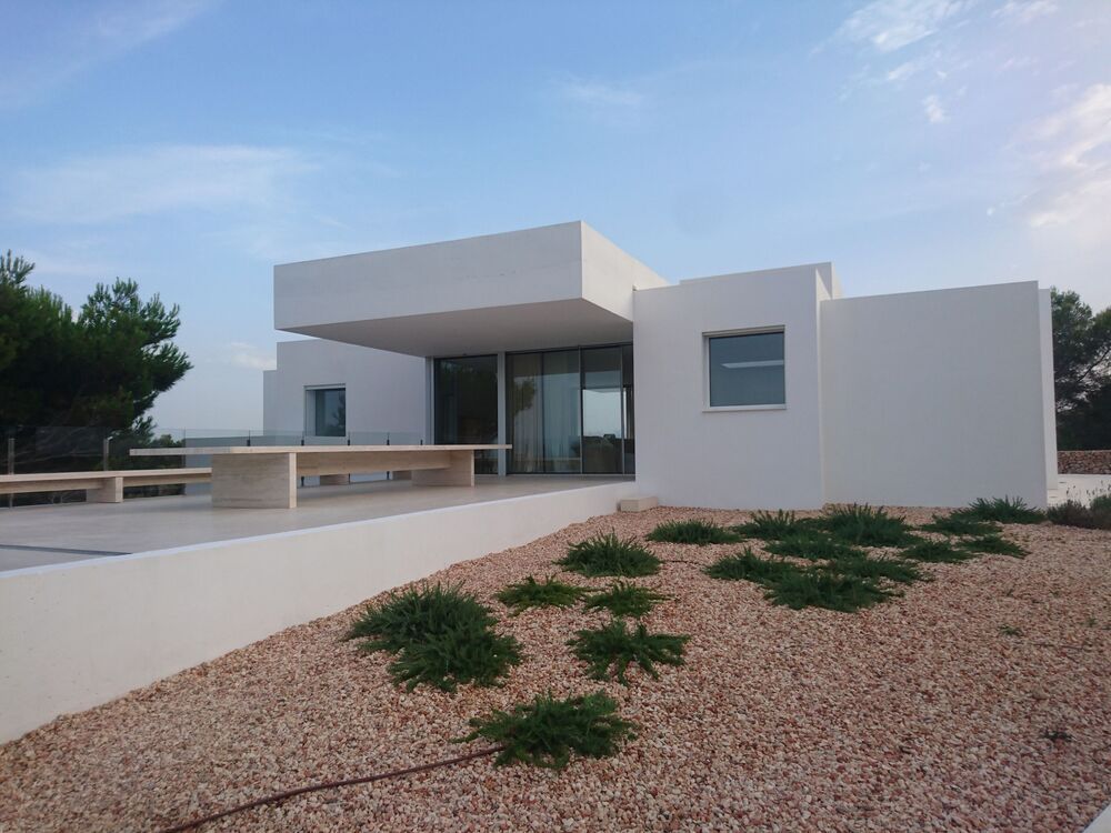 Casa blanca moderna con techo plano, grandes ventanales y zona de estar al aire libre en un día soleado.
