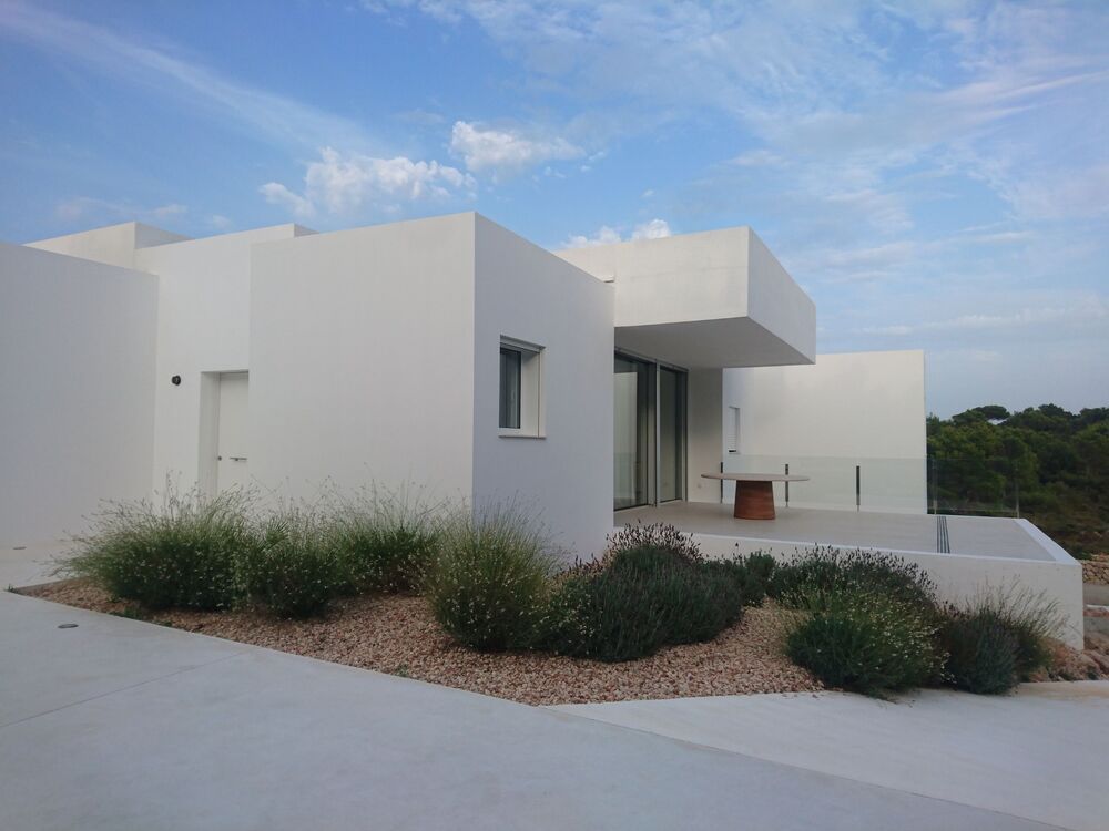 Casa blanca moderna con techo plano, patio y jardinería contra un cielo azul.