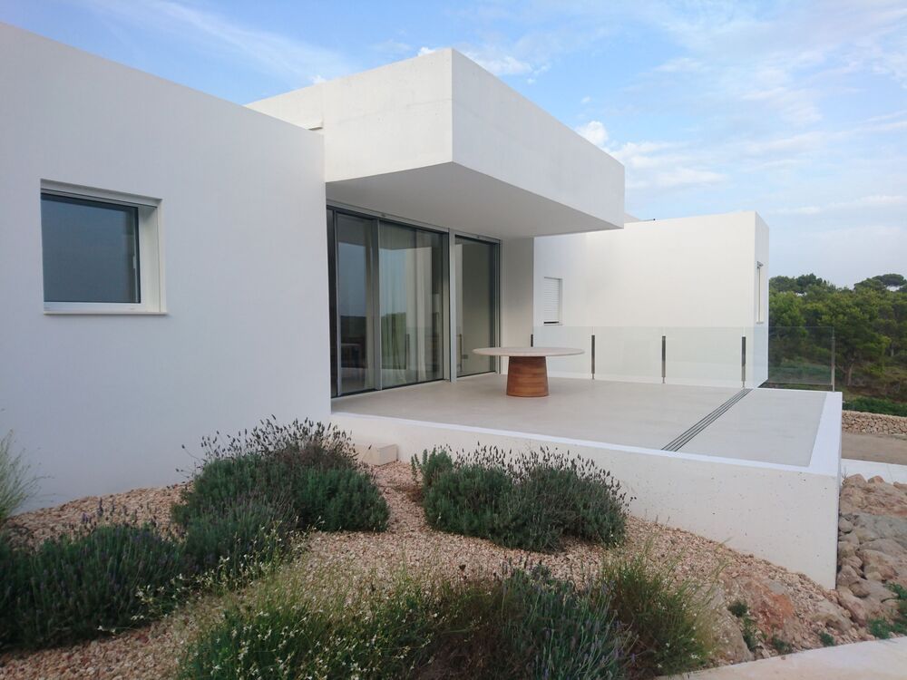Casa blanca moderna con patio, plantas en primer plano y barandilla de vidrio transparente.