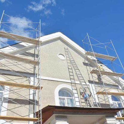Hombre en una escalera pintando la fachada de una casa, con andamios a ambos lados, contra un cielo azul.