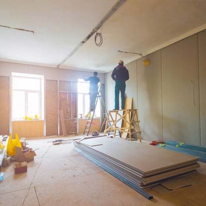 Dos trabajadores instalan paneles de yeso en una habitación en construcción. Se ven andamios, materiales y una ventana.