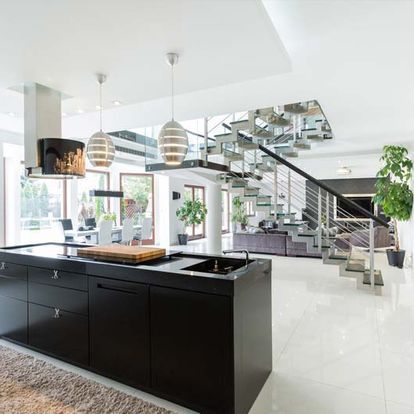 Cocina moderna con isla negra, paredes blancas, escalera de cristal y lámparas colgantes.