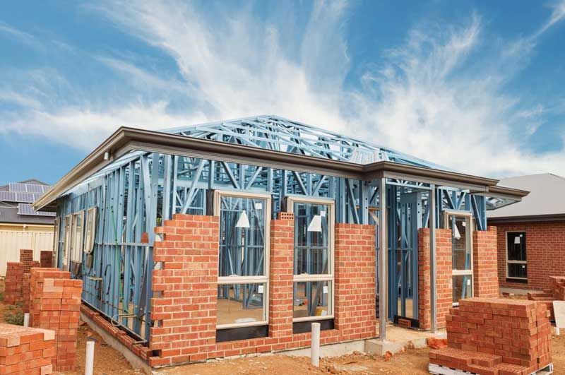 Casa en construcción: estructura de metal, fachada parcialmente de ladrillo, cielo azul.