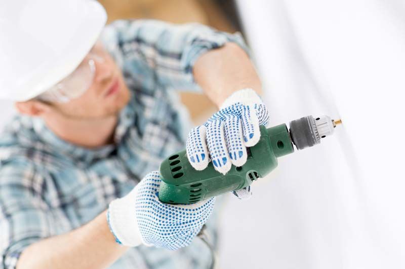 Una persona que usa gafas de seguridad y casco perfora una superficie blanca, sosteniendo un taladro eléctrico verde con guantes azules.