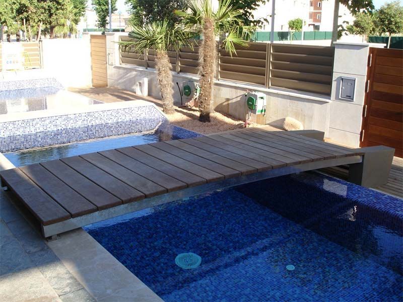 Terraza de madera que cubre una piscina de azulejos azules. Palmeras y una valla al fondo.