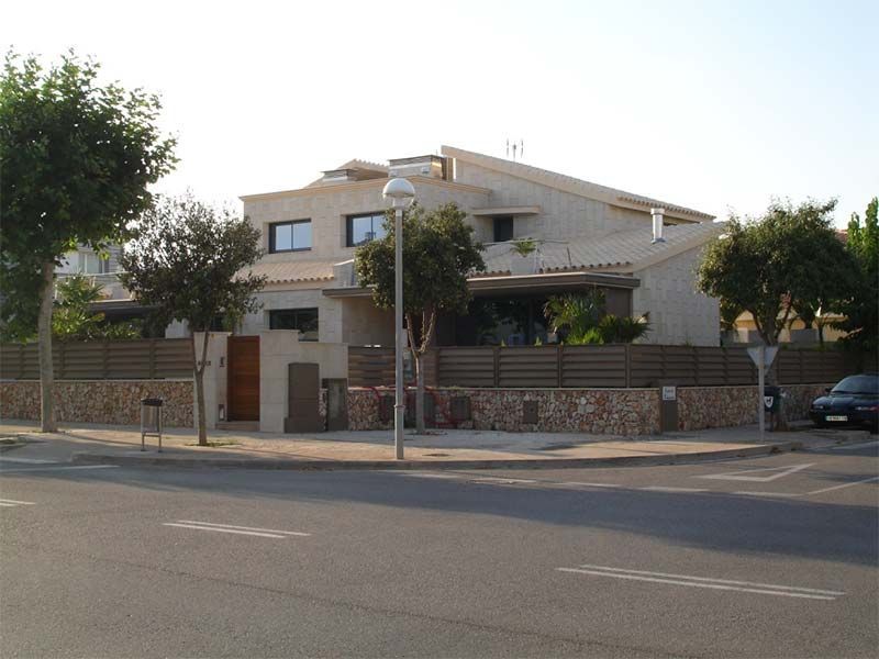Casa de piedra beige con valla baja de color marrón, árboles y calle.