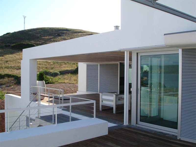 Casa blanca moderna con patio exterior, terraza de madera y puerta corrediza de vidrio. Vistas a la ladera.