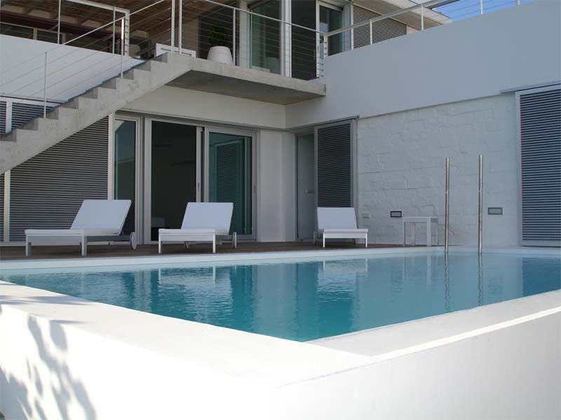 Piscina con tumbonas blancas, escaleras y un moderno edificio blanco.