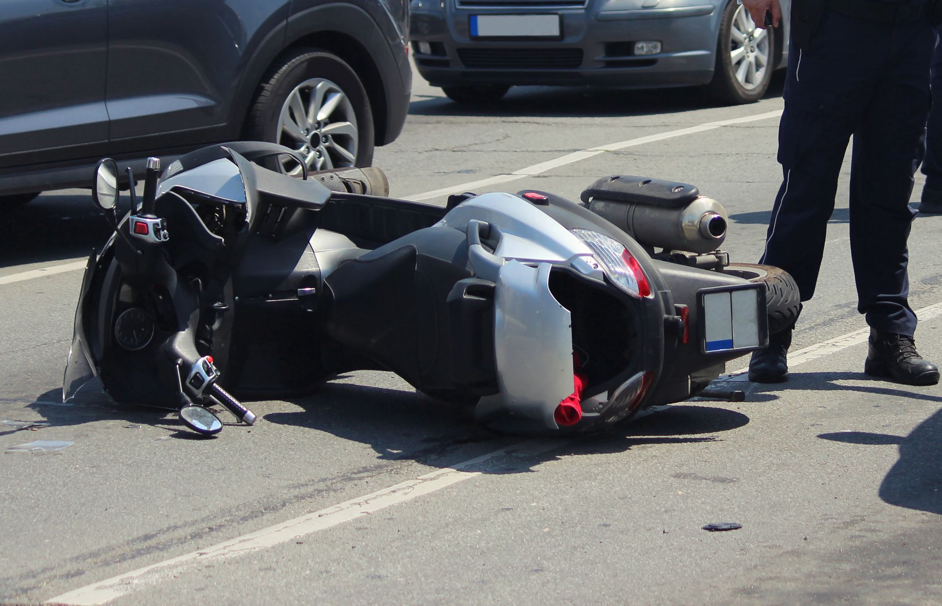 Un scooter renversé sur une route, des voitures stationnées à proximité et les jambes d'une personne debout visibles