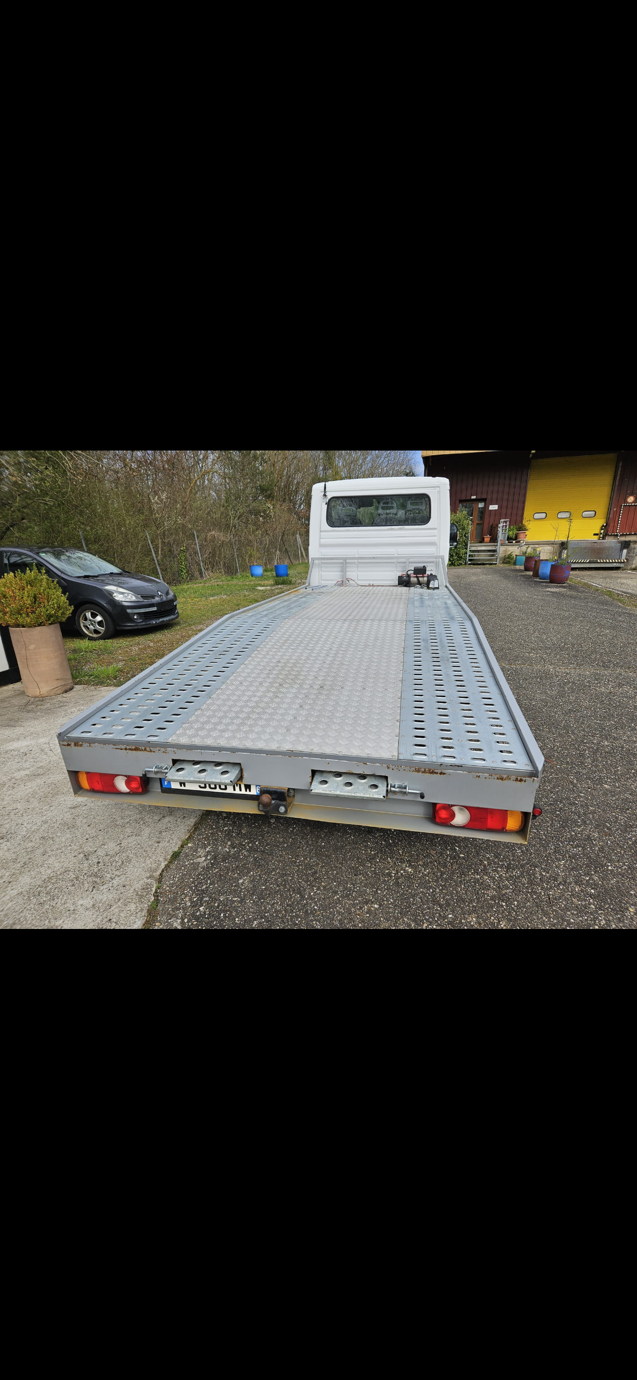 Camion de dépannage gris à plateau stationné sur du gravier, vu de l'arrière, avec une cabine blanche et un bâtiment jaune en arrière-plan
