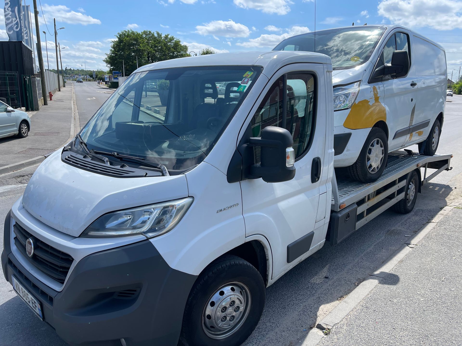 Un camion de dépannage blanc à plateau transporte une petite camionnette blanche dans une rue ensoleillée