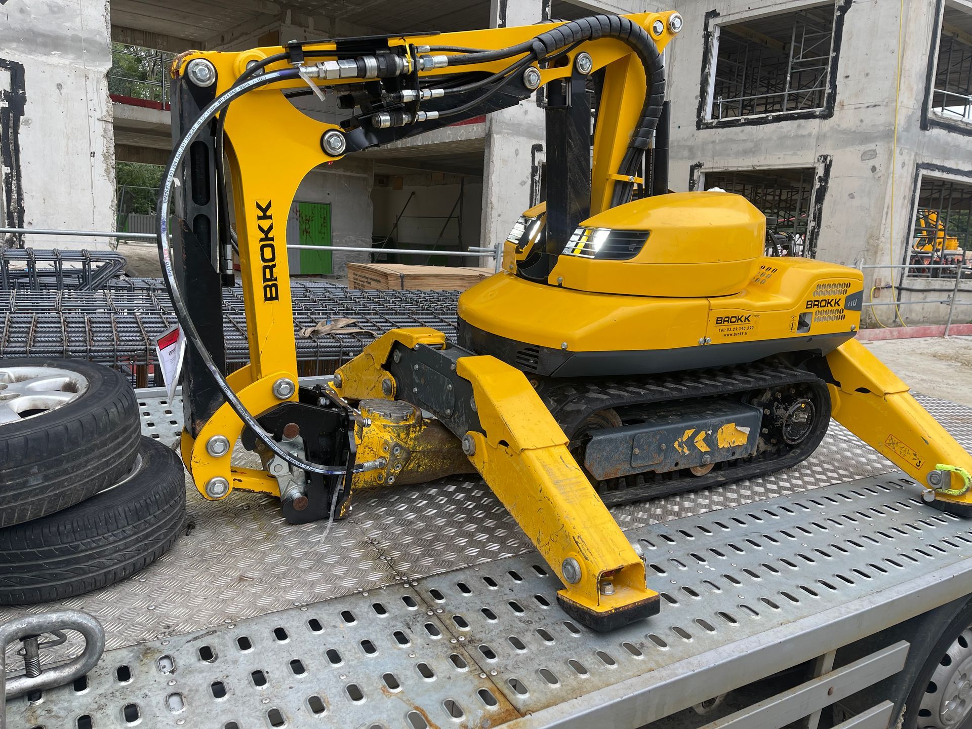 Minipelle chenillée jaune sur une plateforme métallique d'une remorqueuse, bâtiments en construction en arrière-plan