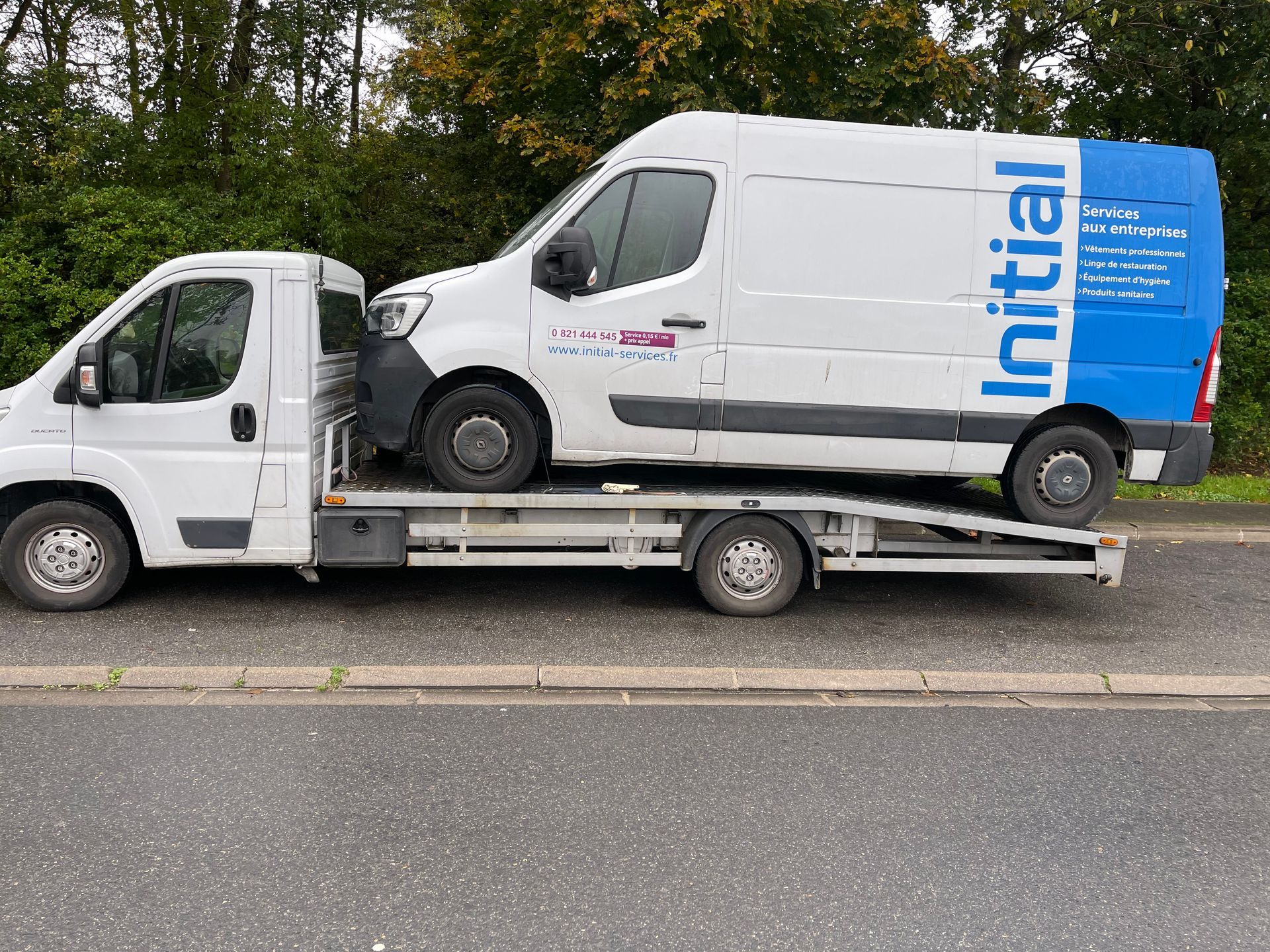 Une camionnette blanche chargée sur une dépanneuse à plateau, garée au bord d'une route avec des arbres en arrière-plan