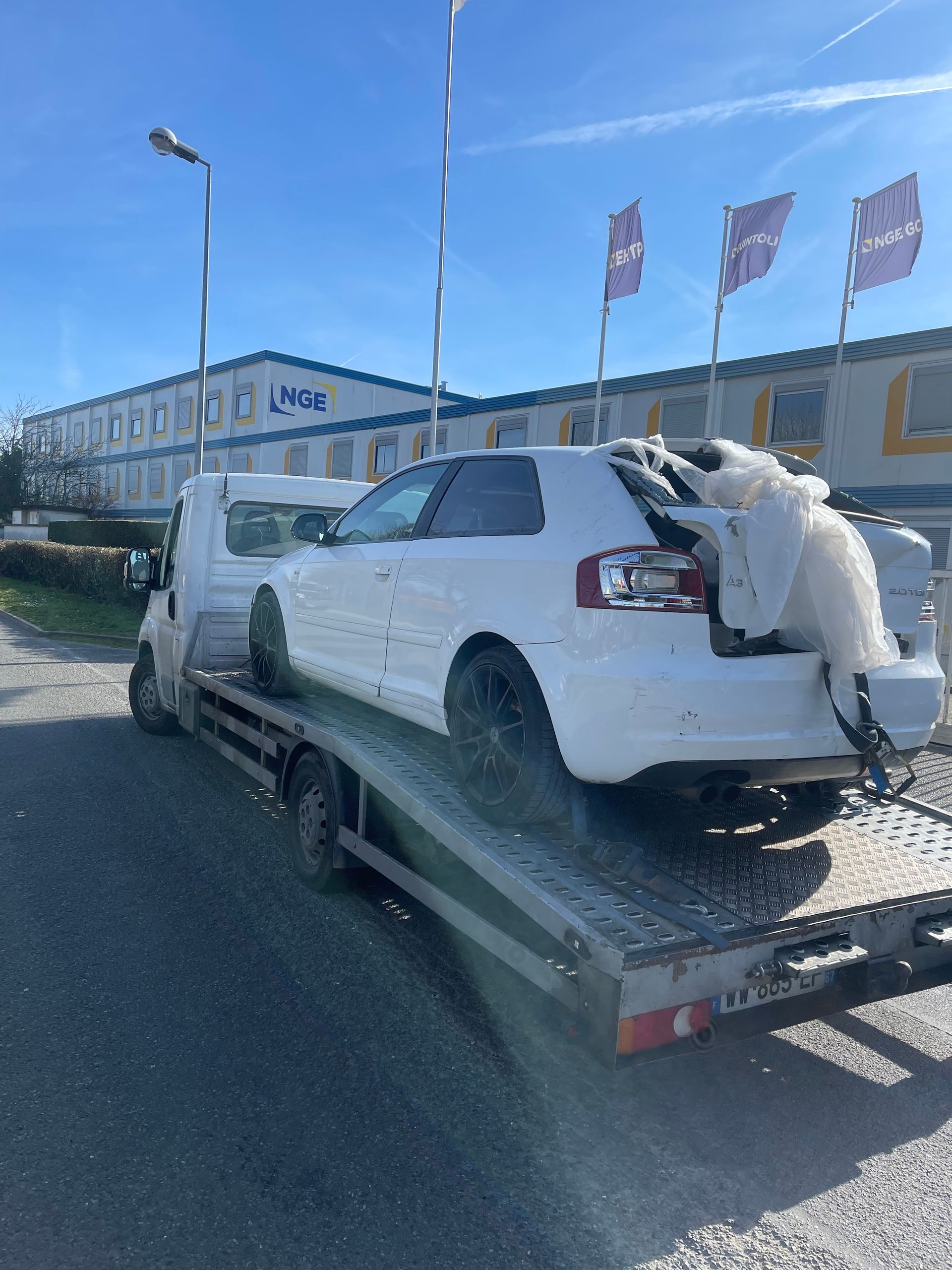 Un voiture blanche accidentée sur le plateau d'une dépanneuse devant un bâtiment avec des drapeaux en arrière-plan