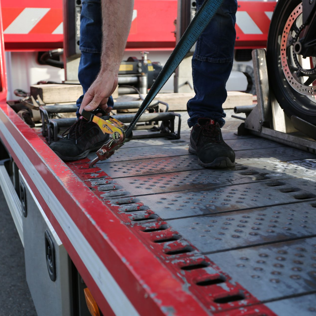 Un ouvrier charge une moto sur une remorque rouge à l'aide d'une sangle