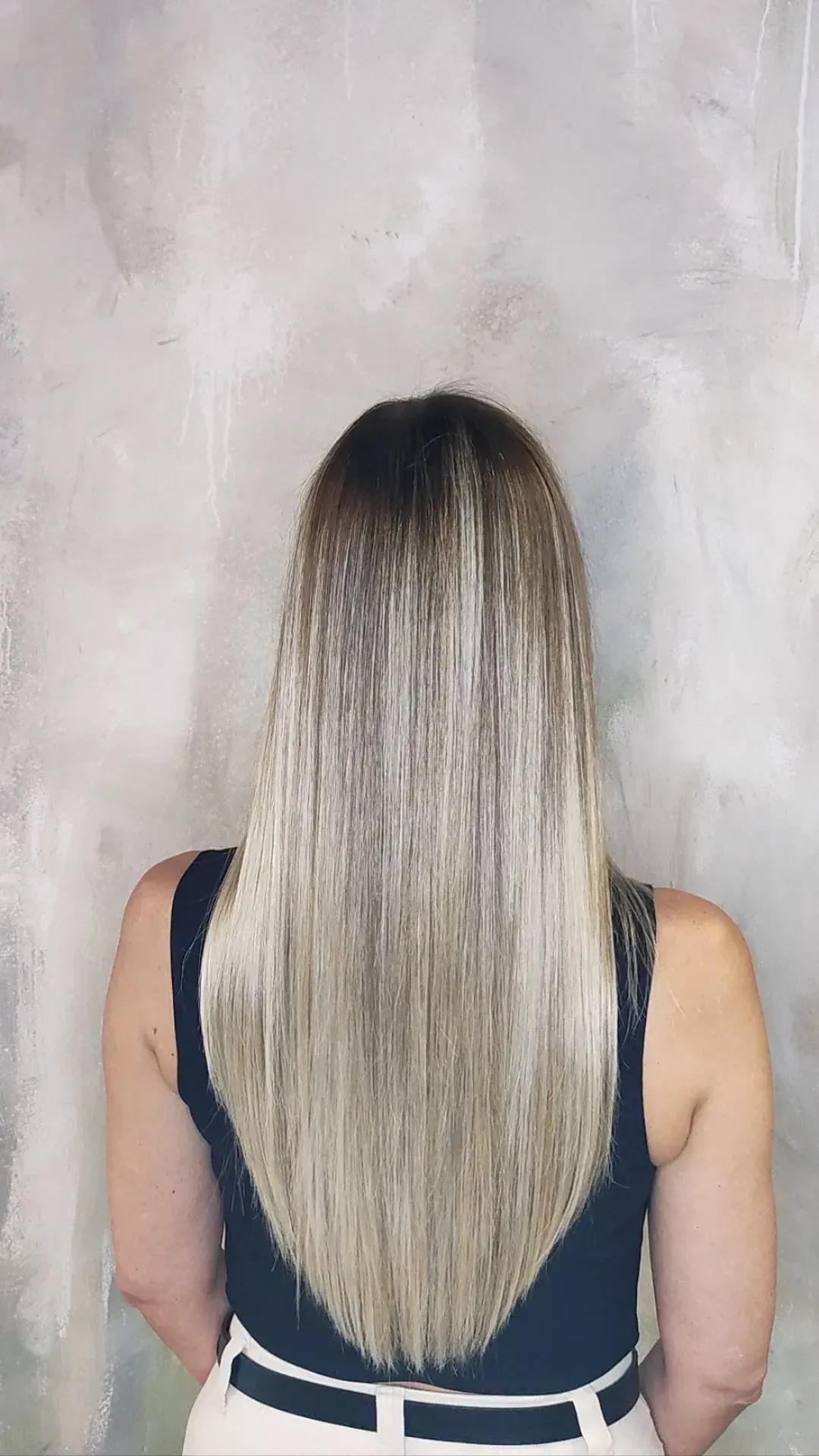 Vista trasera de una persona con cabello largo rubio degradado contra una pared gris.