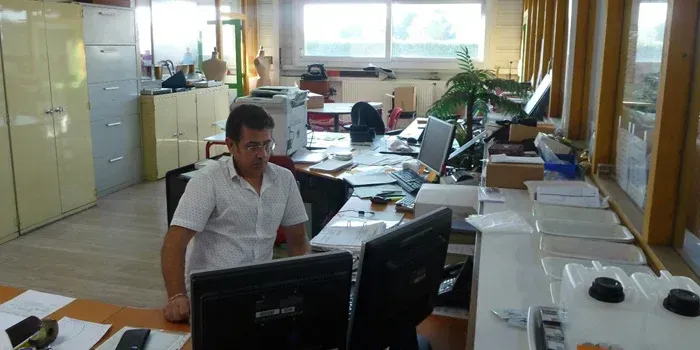 Un homme est assis devant un ordinateur dans un bureau en désordre. Il porte une chemise blanche et regarde l'écran. La lumière du soleil entre par la fenêtre.