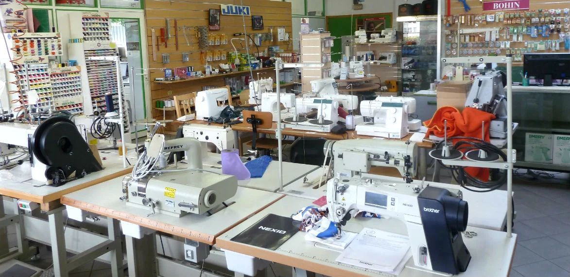 Des machines à coudre sont exposées sur des tables à l'intérieur d'un magasin, avec des étagères remplies de fournitures en arrière-plan.