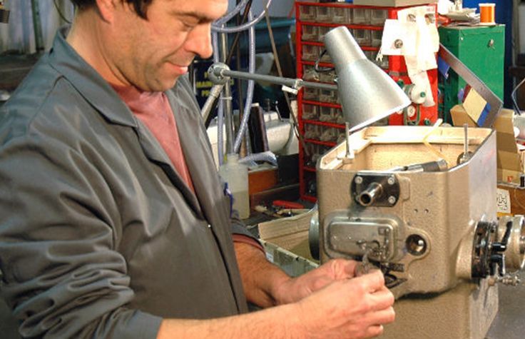 Un homme travaille sur des machines, concentré, vêtu d'une blouse de laboratoire grise, éclairé par une lampe dans un atelier.