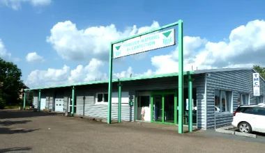 Extérieur d'un bâtiment commercial gris avec une enseigne et des finitions vertes, voiture blanche garée, ciel bleu.
