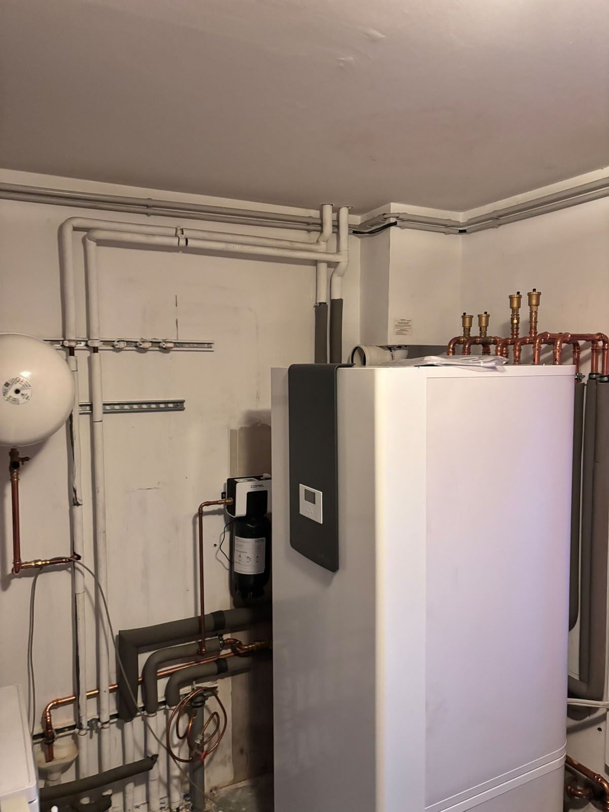 Heizungsanlage mit Rohrleitungen, weißem Tank und wandmontierten Komponenten in einem Hauswirtschaftsraum.