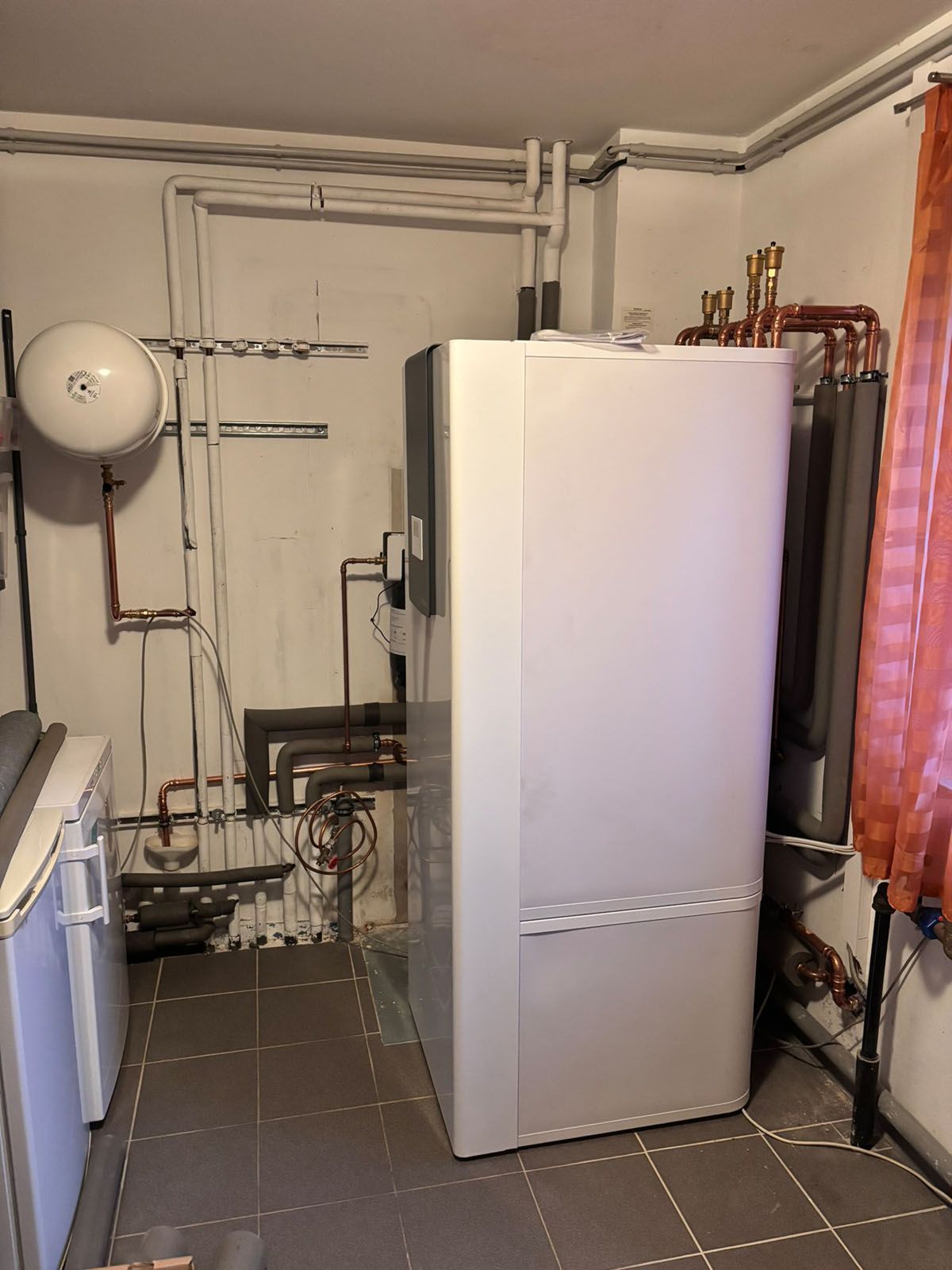 Weißer Ofen in einem Hauswirtschaftsraum mit Rohren, einem kleinen Gerät und einem Fenster mit Vorhang.