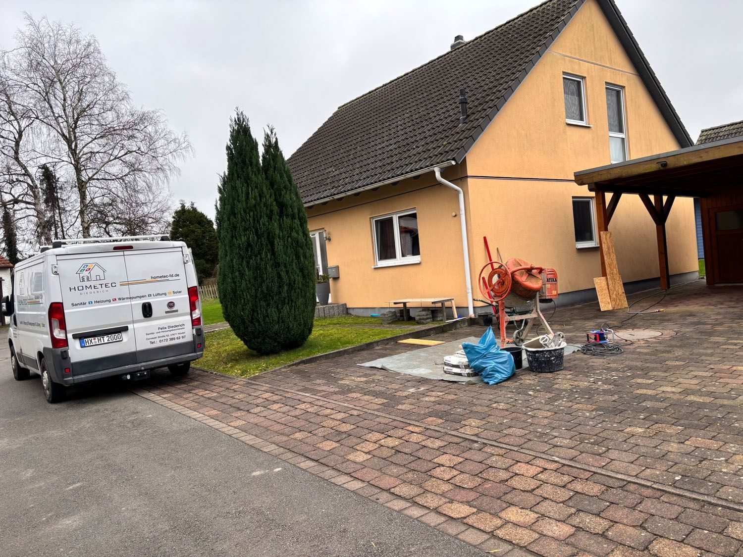 Ein gelbes Haus mit Baumaschinen, ein weißer Lieferwagen und ein grüner Busch an einem bewölkten Tag.