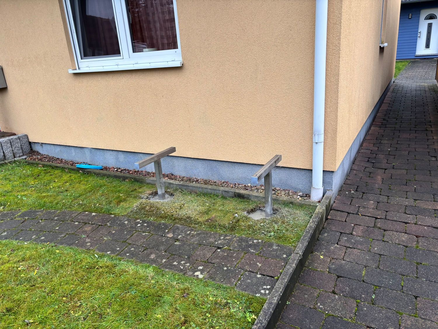 Außenansicht eines Gebäudes mit einer gelben Stuckwand, einem Bürgersteig und zwei Metallpfosten im Gras.