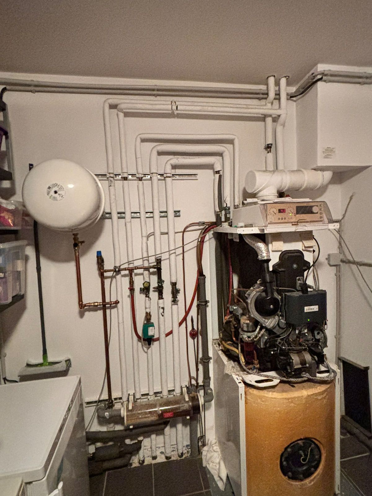 Ofen und Sanitäranlagen in einem Hauswirtschaftsraum mit Rohren, Ventilen und einem weißen Ausdehnungsgefäß.