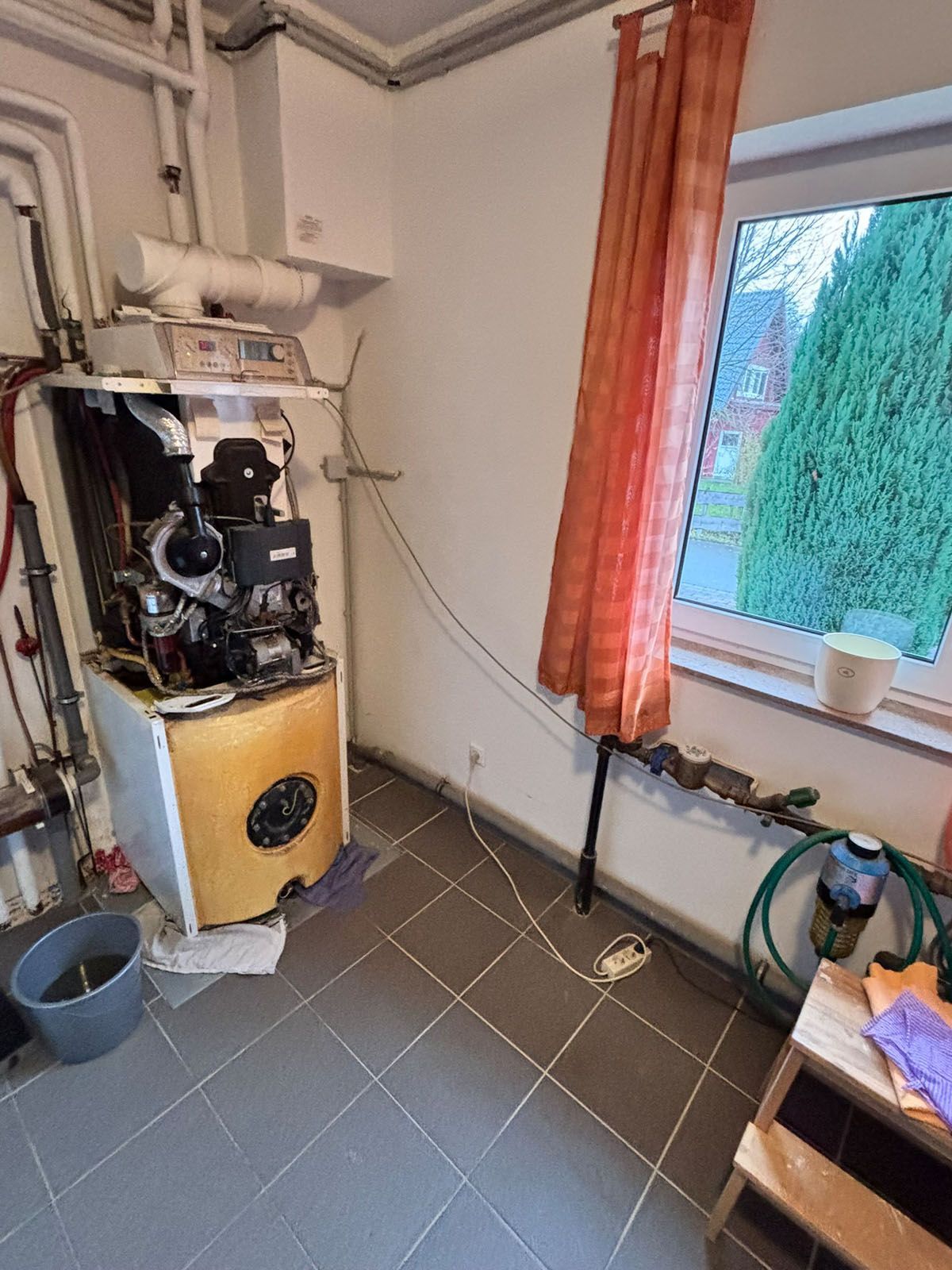 Ein Heizraum mit einem Gaskessel, einem Fenster mit Vorhang und Haushaltsgegenständen.