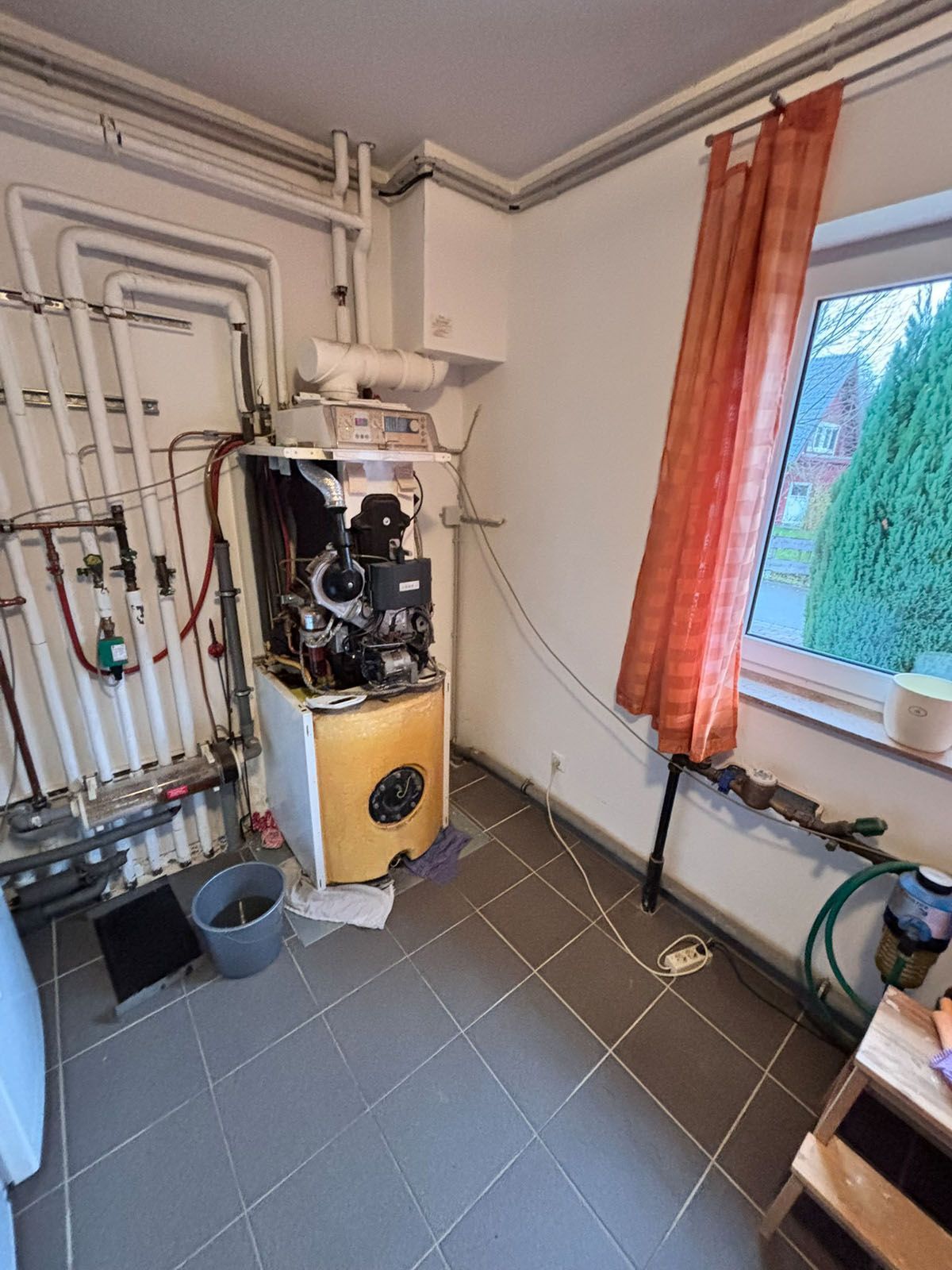 Ein Hauswirtschaftsraum mit einem Heizkessel, Rohren und einem Fenster mit einem roten Vorhang.