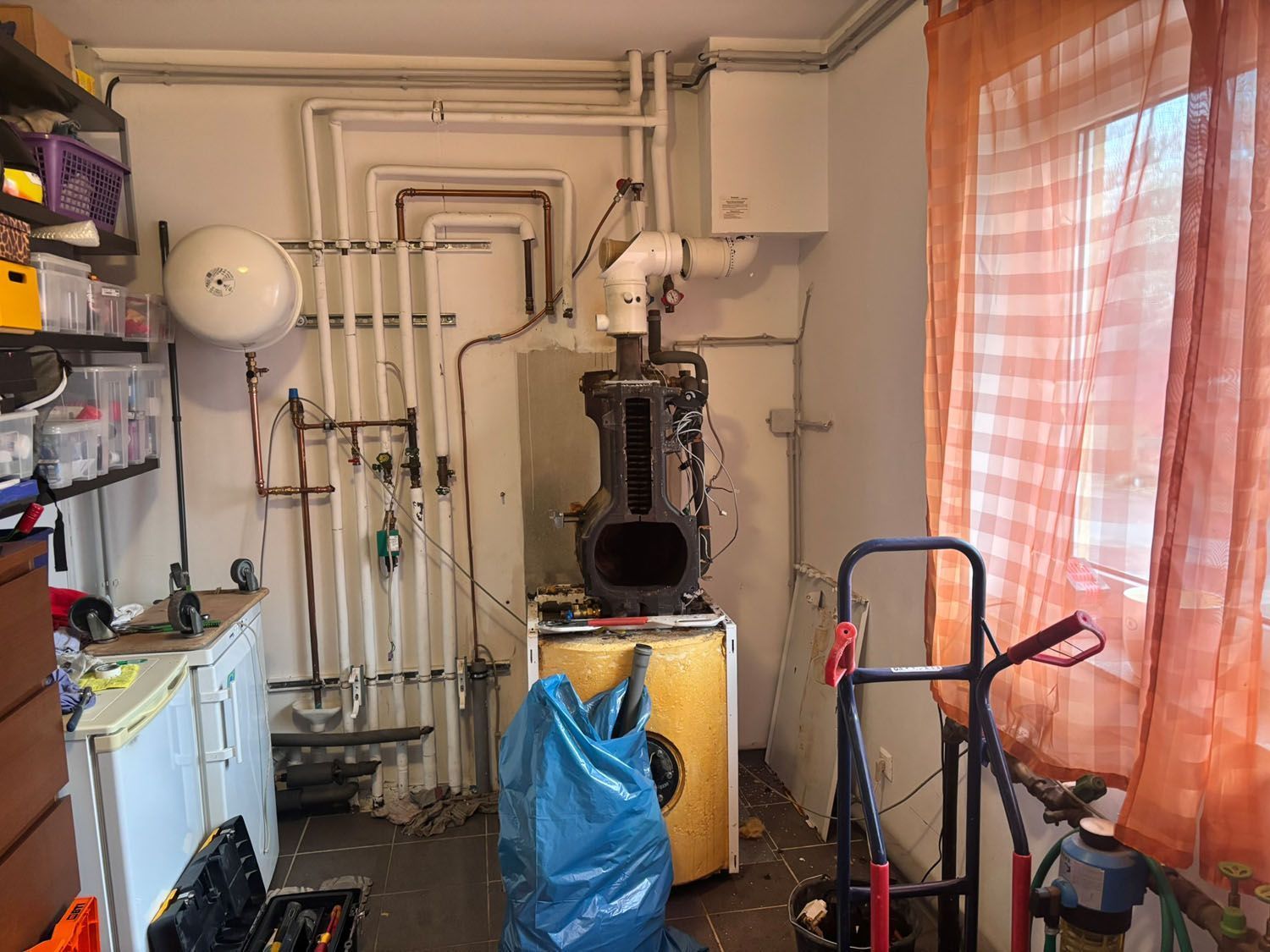 Ein überfüllter Hauswirtschaftsraum mit Heizgeräten, Werkzeugen und einem Fenster mit orangefarbenen Vorhängen.