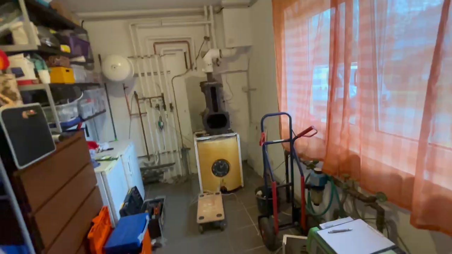 Unordentlicher Keller: braune Kommode, Lagerregale, alter Ofen, Fenster mit orangefarbenen Vorhängen.