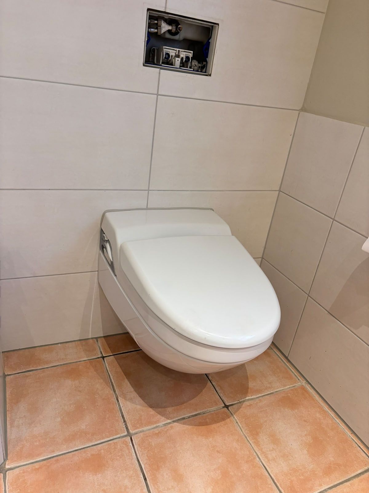 Wandmontierte Toilette in einem Badezimmer mit beigen Fliesen. Leere Öffnung für den Spülmechanismus.