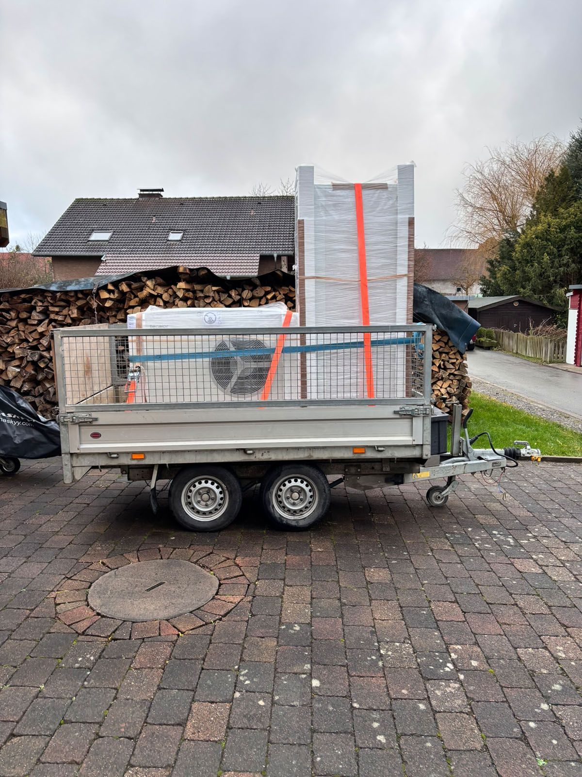 Ein mit Baumaterialien beladener und mit Gurten gesicherter Anhänger steht auf einer gepflasterten Auffahrt.