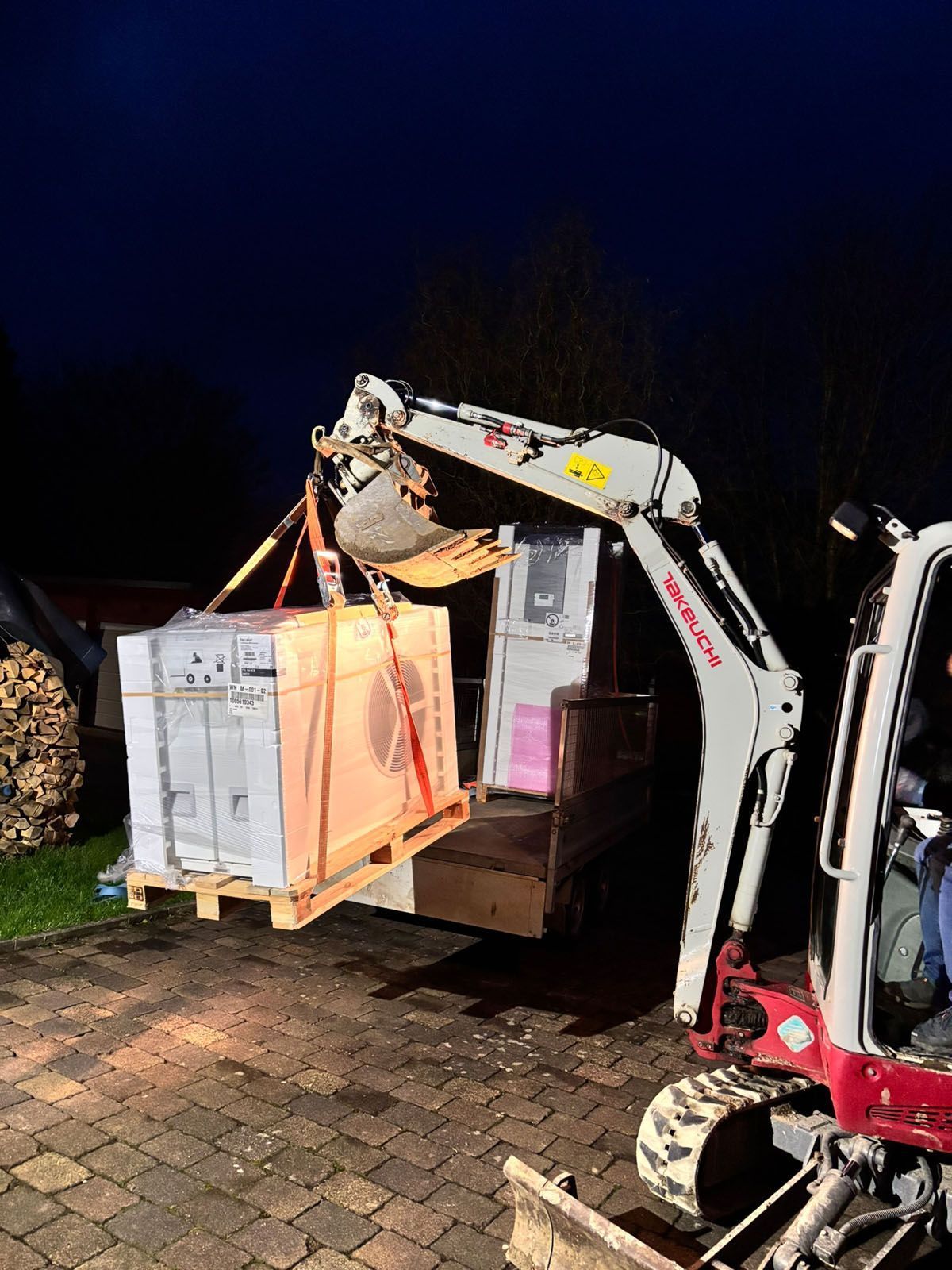 Minibagger hebt nachts eine Palette mit Kisten auf einen LKW.