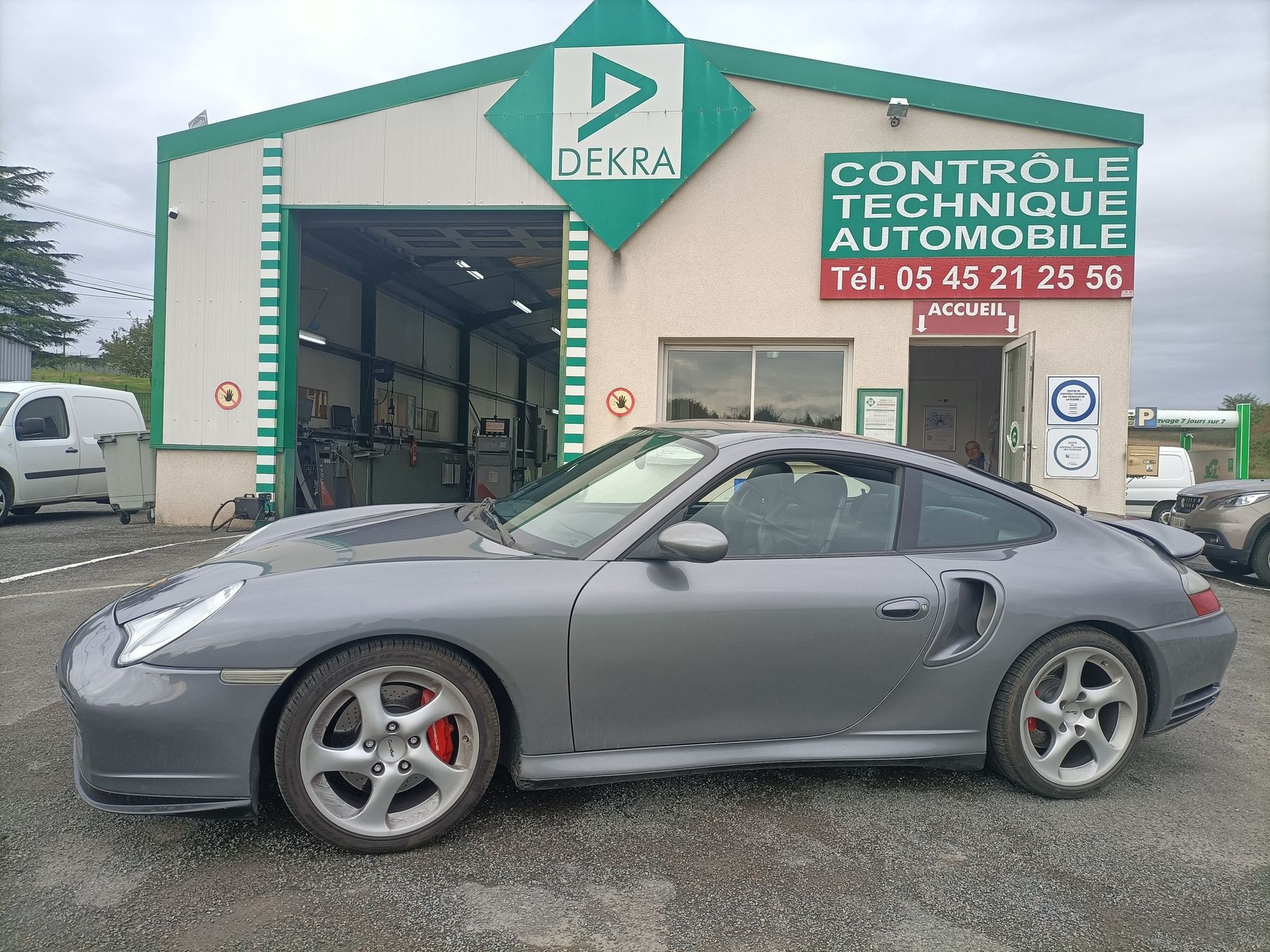 Une Porsche grise est garée devant le centre de contrôle technique automobile DEKRA.