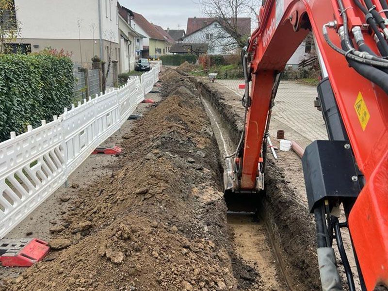 Ein roter Bagger gräbt neben einem weißen Zaun einen Graben in die Erde.