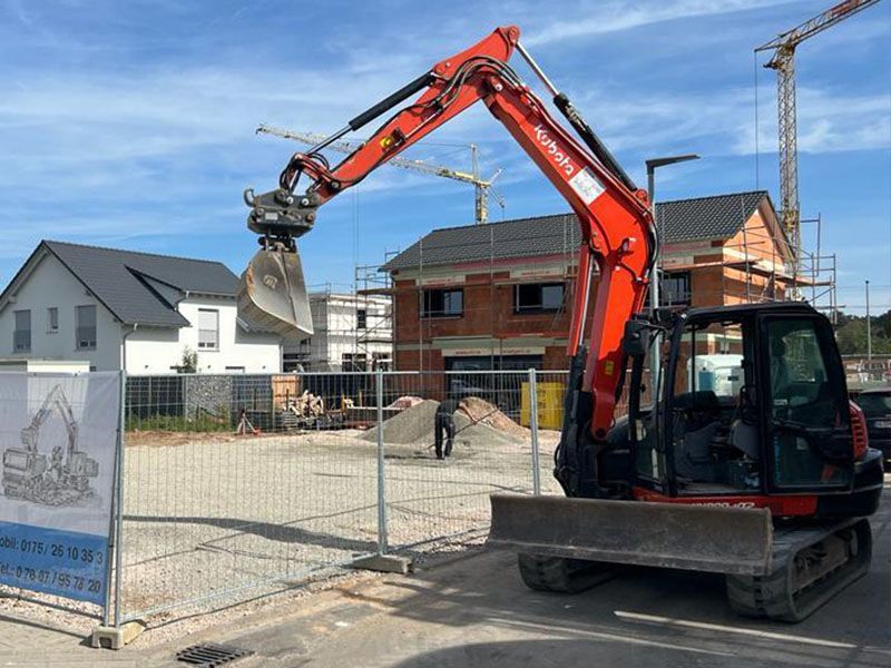 Ein orangefarbener Bagger steht vor einem Zaun auf einer Baustelle.