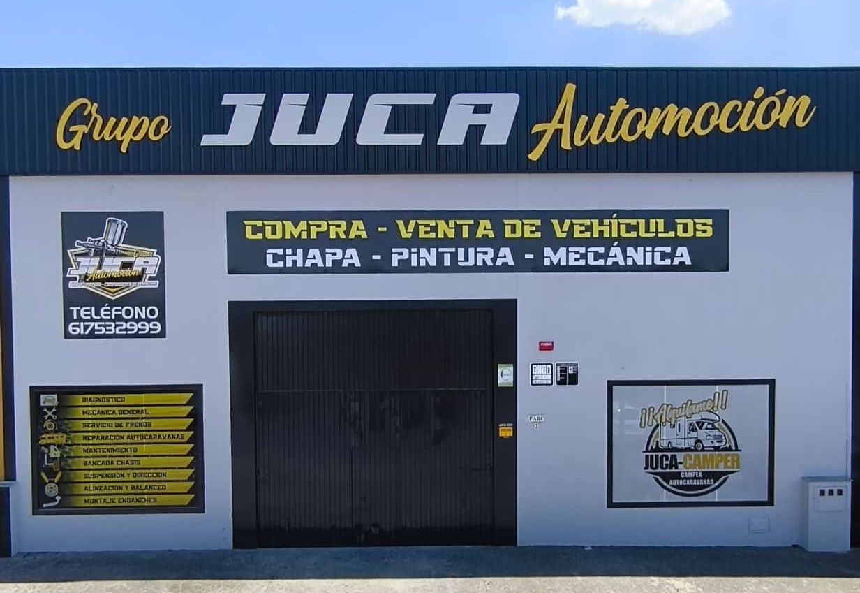 Fachada del taller Juca Automoción, con un letrero azul y amarillo, una puerta de garaje negra y anuncios de servicios.