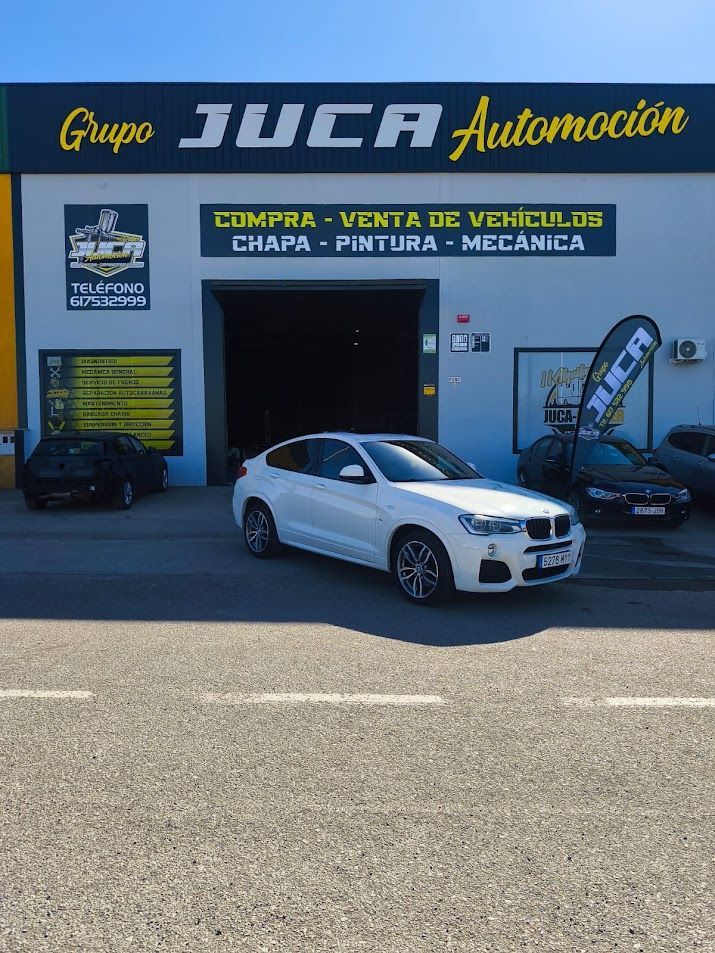 Sedán blanco aparcado frente al taller de coches JUCA Automoción, con zona de servicio y letreros.