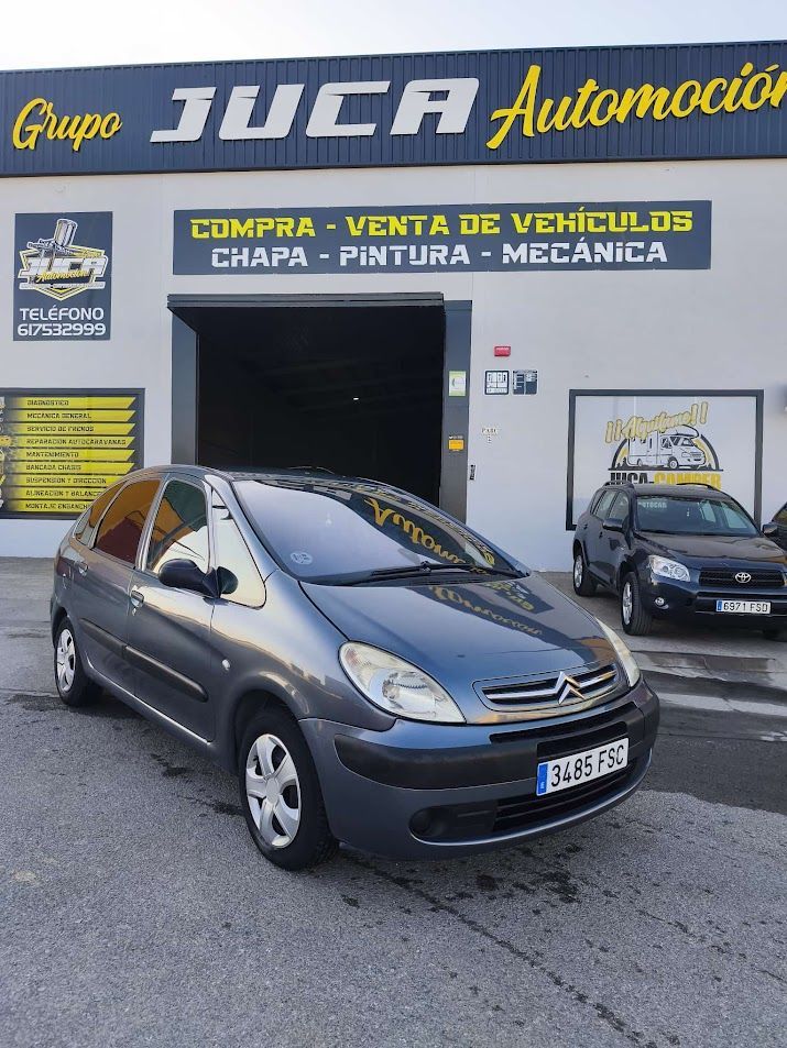 Un coche gris aparcado delante de un concesionario de coches usados y taller de reparación de automóviles con un gran letrero de “Tuca Automoción”.