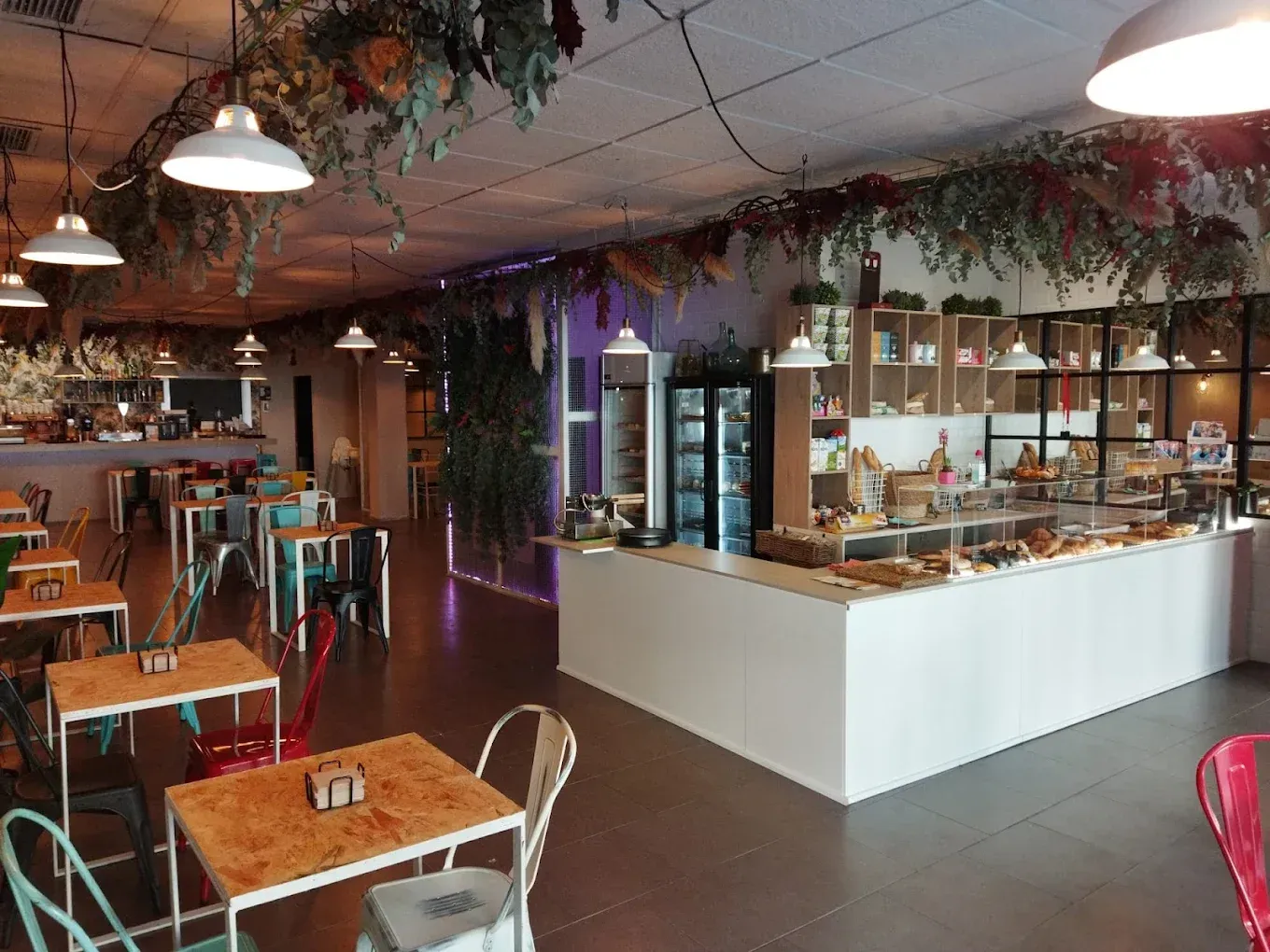 Un acogedor interior de cafetería con mesas con tablero de madera, sillas de metal de colores, plantas colgantes y un mostrador con pasteles.